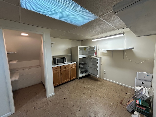 kitchen with baseboards, a drop ceiling, stainless steel microwave, brown cabinets, and light floors