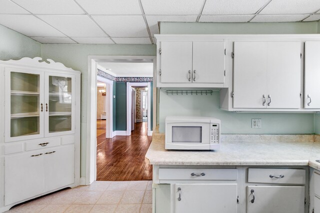 kitchen with white microwave, a drop ceiling, light countertops, light tile patterned floors, and white cabinets