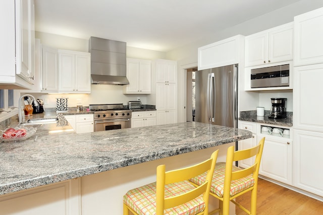 kitchen featuring wall chimney exhaust hood, a kitchen breakfast bar, stainless steel appliances, light stone countertops, and white cabinets