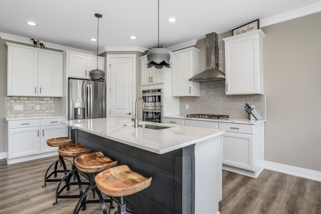 kitchen with wall chimney exhaust hood, hanging light fixtures, stainless steel appliances, wood-type flooring, and a kitchen island with sink