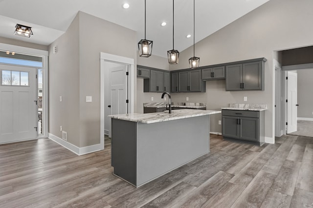 kitchen featuring wood-type flooring, decorative light fixtures, a center island with sink, gray cabinets, and light stone countertops