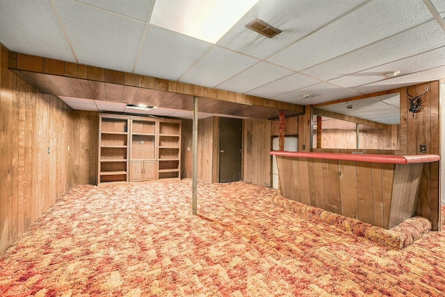 basement with carpet flooring, a paneled ceiling, bar, and wooden walls