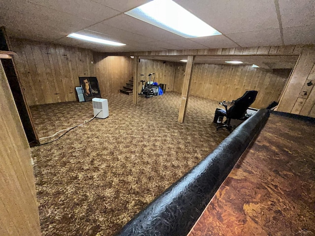 basement featuring dark colored carpet, a paneled ceiling, and wooden walls