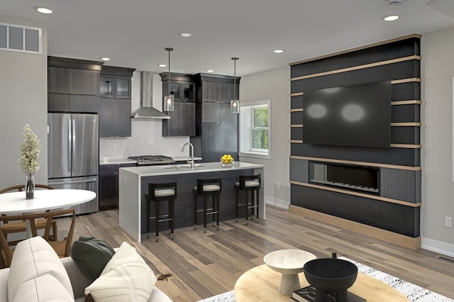 kitchen with stainless steel fridge, wall chimney exhaust hood, a kitchen island with sink, sink, and pendant lighting