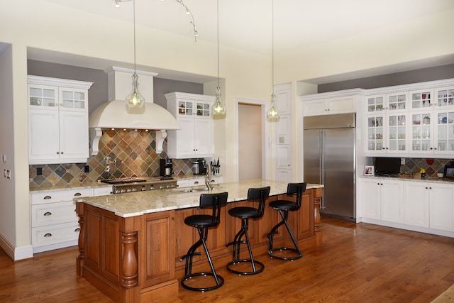 kitchen featuring an island with sink, glass insert cabinets, dark wood-type flooring, hanging light fixtures, and stainless steel built in fridge