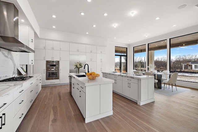 kitchen featuring white cabinetry, wall chimney exhaust hood, stainless steel appliances, tasteful backsplash, and a center island with sink