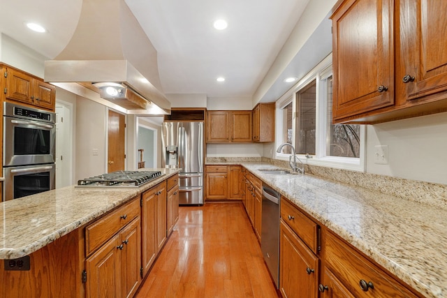 kitchen with island exhaust hood, appliances with stainless steel finishes, light wood-type flooring, light stone counters, and sink