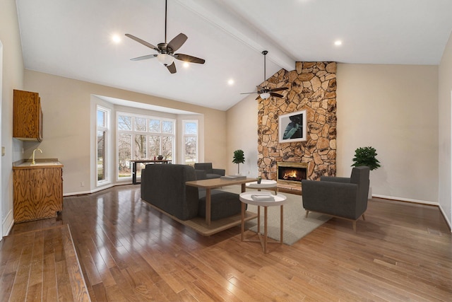 living room featuring ceiling fan, sink, a stone fireplace, vaulted ceiling with beams, and hardwood / wood-style floors