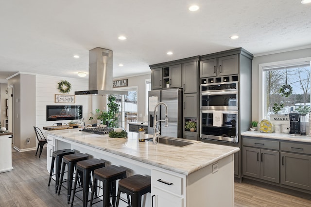 kitchen with island exhaust hood, an island with sink, gray cabinets, a breakfast bar, and appliances with stainless steel finishes