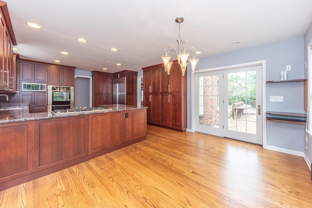 kitchen with an inviting chandelier, light hardwood / wood-style flooring, built in appliances, dark stone counters, and pendant lighting