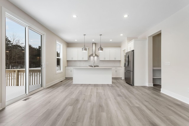 kitchen featuring wall chimney range hood, hanging light fixtures, stainless steel fridge with ice dispenser, an island with sink, and white cabinetry