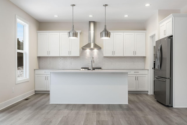 kitchen with stainless steel fridge, wall chimney exhaust hood, a kitchen island with sink, decorative light fixtures, and white cabinetry