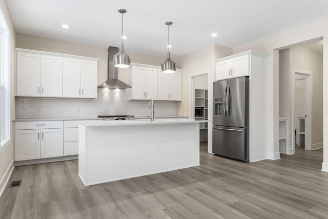 kitchen featuring appliances with stainless steel finishes, wall chimney range hood, decorative light fixtures, a center island with sink, and white cabinets