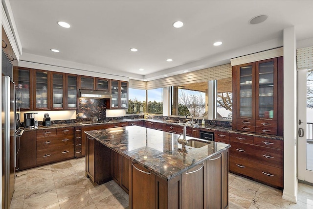 kitchen featuring an island with sink, stainless steel gas cooktop, dark stone countertops, dark brown cabinetry, and sink