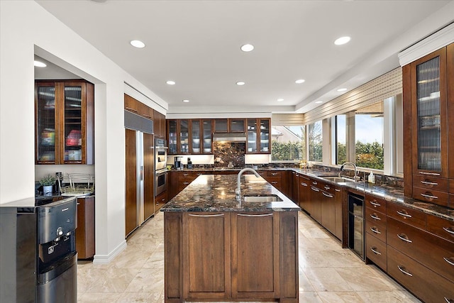 kitchen with wine cooler, a kitchen island with sink, dark stone counters, and sink