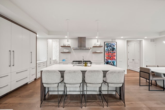 kitchen with sink, hanging light fixtures, wall chimney range hood, light hardwood / wood-style flooring, and an island with sink