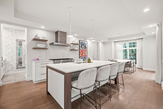 kitchen with white cabinetry, sink, wall chimney exhaust hood, light hardwood / wood-style floors, and a center island with sink