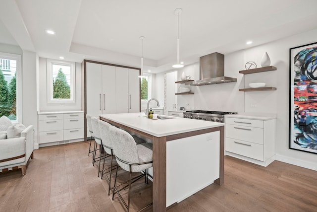 kitchen with a center island with sink, wall chimney range hood, sink, hanging light fixtures, and white cabinetry