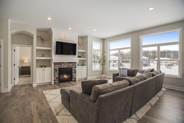 living room with built in shelves, a stone fireplace, and dark wood-type flooring