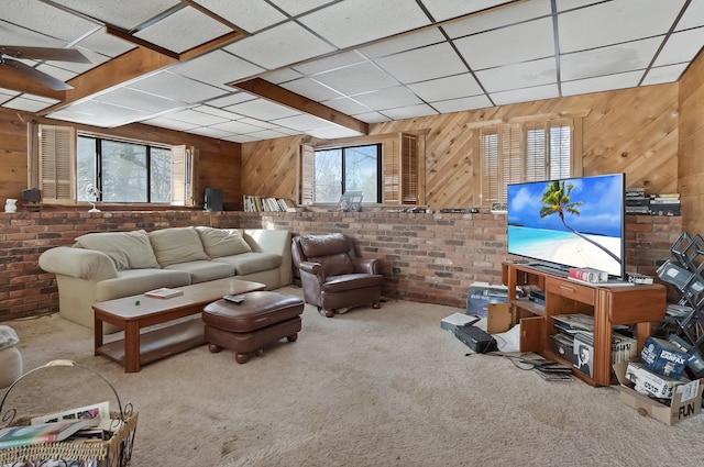 carpeted living room with a drop ceiling, wood walls, and brick wall