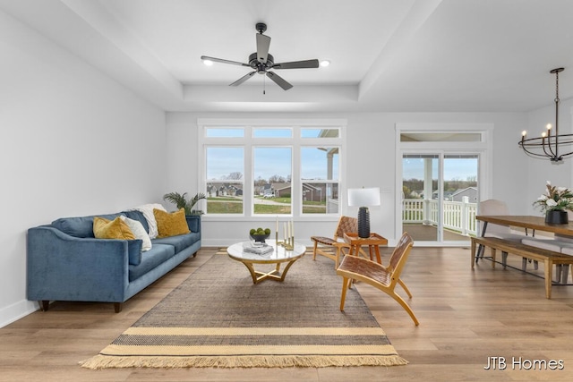 living area featuring baseboards, light wood-style flooring, a raised ceiling, and ceiling fan with notable chandelier
