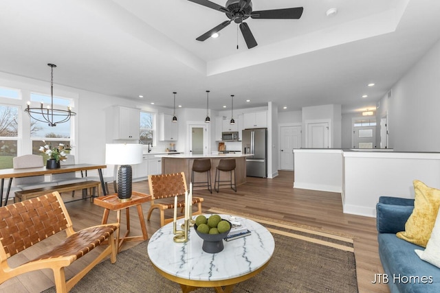 living area featuring light wood-style floors, a raised ceiling, ceiling fan with notable chandelier, and recessed lighting