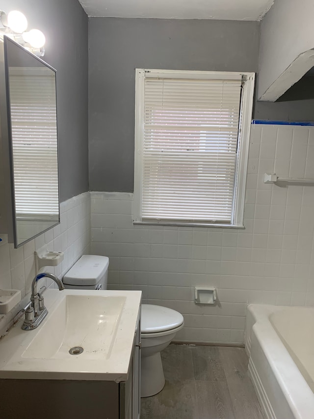 bathroom with a tub to relax in, wood-type flooring, toilet, vanity, and tile walls