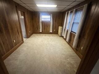 basement with a paneled ceiling, radiator heating unit, wooden walls, and light carpet