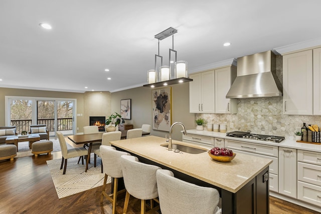 kitchen with pendant lighting, stainless steel gas stovetop, wall chimney range hood, sink, and an island with sink