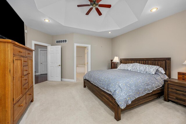 bedroom with ceiling fan, light colored carpet, a tray ceiling, and ensuite bath