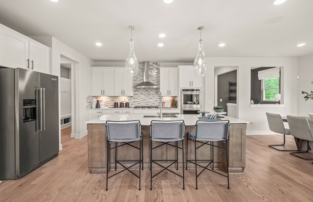 kitchen featuring a center island with sink, white cabinets, wall chimney range hood, light hardwood / wood-style flooring, and stainless steel appliances