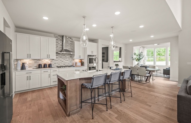 kitchen with stainless steel appliances, a kitchen island with sink, wall chimney range hood, pendant lighting, and white cabinets