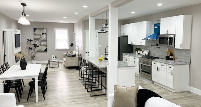 kitchen with wall chimney exhaust hood, light stone countertops, appliances with stainless steel finishes, decorative light fixtures, and white cabinetry