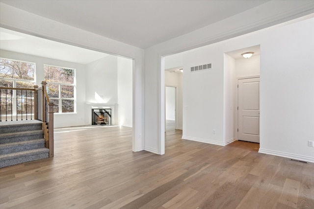 unfurnished living room featuring visible vents, light wood-style flooring, stairway, a warm lit fireplace, and baseboards