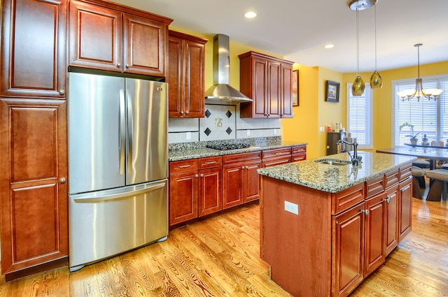 kitchen featuring stainless steel appliances, an island with sink, light stone counters, wall chimney exhaust hood, and sink