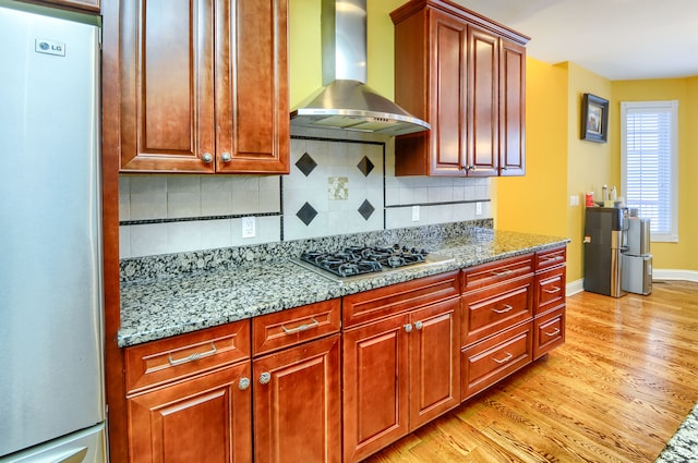 kitchen featuring light stone counters, stainless steel appliances, wall chimney range hood, light hardwood / wood-style floors, and decorative backsplash