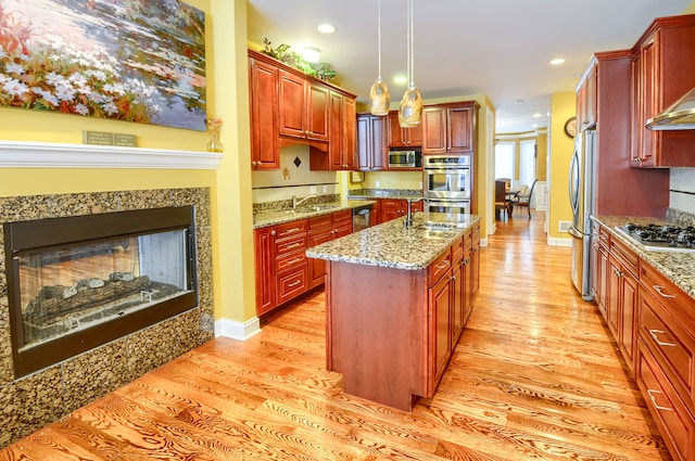 kitchen with light hardwood / wood-style floors, stainless steel appliances, an island with sink, light stone counters, and decorative light fixtures