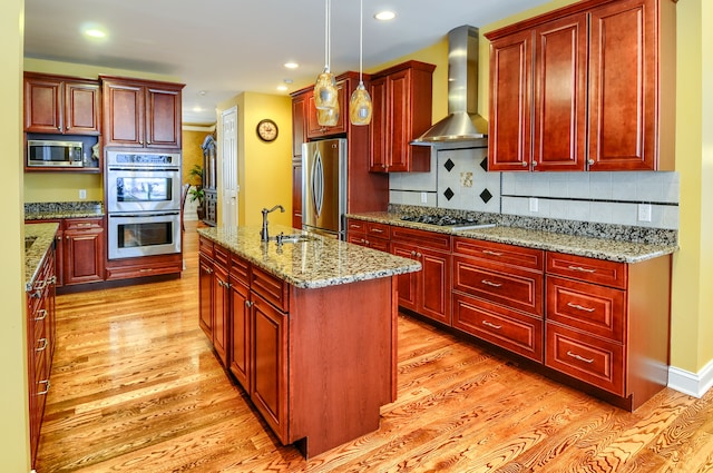 kitchen with stainless steel appliances, an island with sink, light stone countertops, wall chimney range hood, and pendant lighting