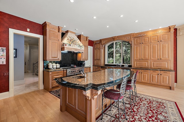 kitchen with backsplash, dark stone countertops, a center island with sink, a kitchen breakfast bar, and custom range hood