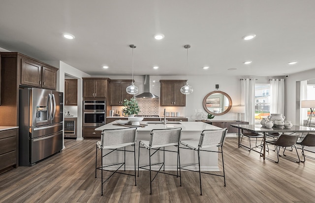 kitchen featuring a center island with sink, appliances with stainless steel finishes, wall chimney range hood, decorative light fixtures, and tasteful backsplash