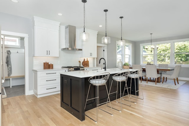 kitchen featuring a kitchen island with sink, light wood-type flooring, wall chimney exhaust hood, white cabinetry, and decorative light fixtures