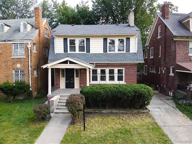 view of front of property featuring brick siding, roof with shingles, covered porch, and a front yard