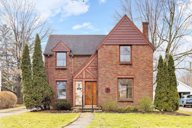 tudor-style house with brick siding, a chimney, a front yard, and a shingled roof