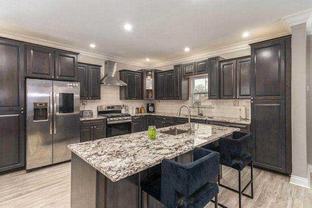 kitchen with wall chimney exhaust hood, a kitchen breakfast bar, light hardwood / wood-style flooring, an island with sink, and appliances with stainless steel finishes