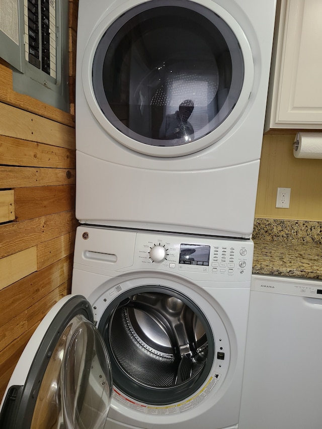 washroom with cabinets, wooden walls, electric panel, and stacked washer / dryer