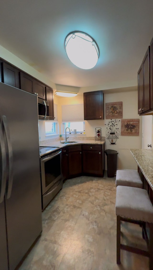 kitchen with sink, appliances with stainless steel finishes, dark brown cabinets, light stone counters, and a kitchen bar