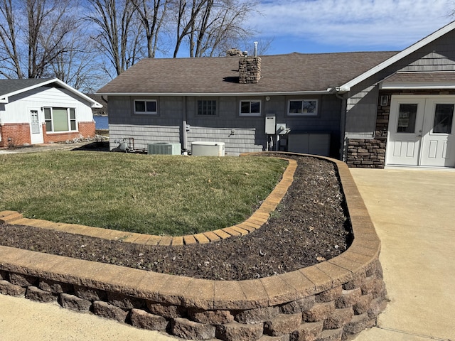 back of house with central AC unit, a shingled roof, concrete driveway, stone siding, and a lawn