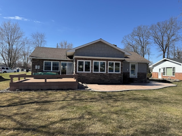 rear view of property featuring a shingled roof, a wooden deck, a lawn, stone siding, and a patio