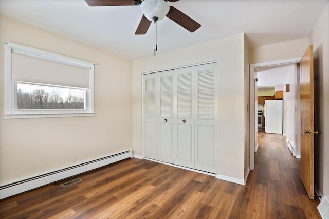 unfurnished bedroom with white fridge, a closet, baseboard heating, dark hardwood / wood-style flooring, and ceiling fan