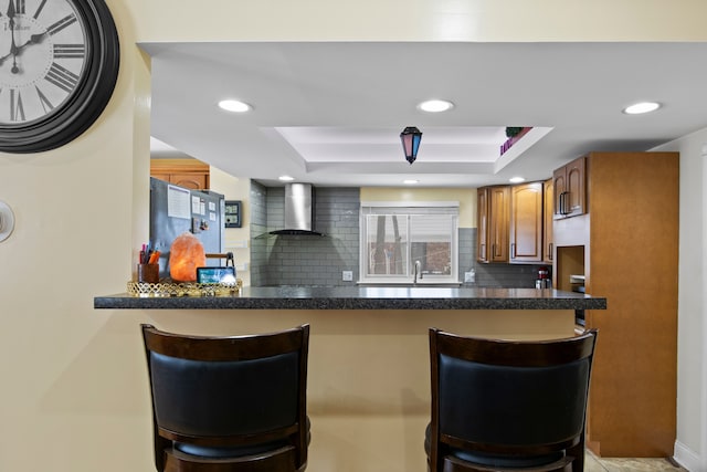 kitchen featuring a breakfast bar area, kitchen peninsula, decorative backsplash, a tray ceiling, and wall chimney range hood